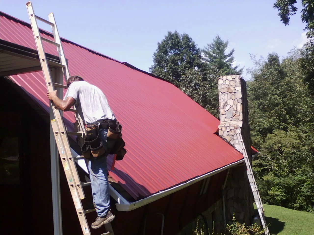 Roofer on a ladder beside a red metal roof and stone chimney — Soffit & Fascia Repair by Roofing Brothers in Norris